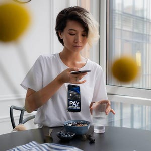 May include: A white t-shirt featuring a graphic print of a smartphone displaying a credit card and the text PAY ATTENTION. The shirt is worn by a person with short dark hair with blonde highlights, sitting at a table with a bowl of berries and a glass.