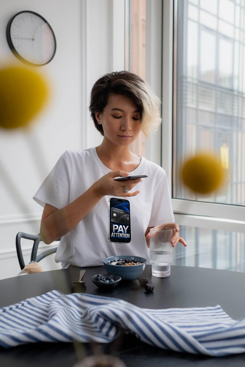 May include: A white t-shirt featuring a graphic print of a smartphone displaying a credit card and the text PAY ATTENTION. The shirt is worn by a person with short dark hair with blonde highlights, sitting at a table with a bowl of berries and a glass.
