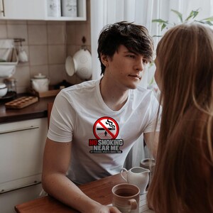 May include: A white short-sleeved t-shirt featuring a circular red no-smoking symbol with a cigarette and the text NO SMOKING NEAR ME MY PERSONAL SPACE IS SMOKE-FREE printed in black and red lettering on the chest area.