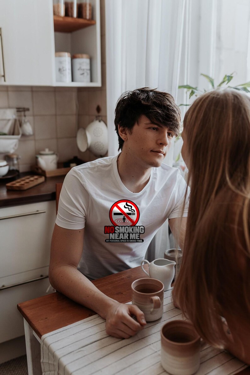 May include: A white short-sleeved t-shirt featuring a circular red no-smoking symbol with a cigarette and the text NO SMOKING NEAR ME MY PERSONAL SPACE IS SMOKE-FREE printed in black and red lettering on the chest area.