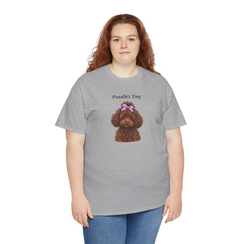 May include: A heather gray short-sleeve t-shirt featuring a graphic of a brown poodle with a purple bow on its head. Above the dog, the text Poodle's Day is printed in black. The shirt has a crew neckline and a standard fit.