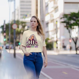 May include: A cream-colored short-sleeve t-shirt featuring a graphic of a woman with long gray hair, the text "HI Ask me anything...", and small icons for new chat, voice, and image, paired with blue denim jeans.