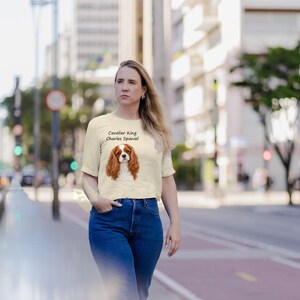 Puede incluir: Una camiseta de manga corta de color crema con una ilustraci&oacute;n gr&aacute;fica de un perro Cavalier King Charles Spaniel y el texto "Cavalier King Charles Spaniel" impreso en la parte delantera, combinada con unos vaqueros azules.