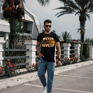 Puede incluir: Una camiseta negra de manga corta con el texto NEVER GIVE UP en letras amarillas sobre una ilustraci&oacute;n gr&aacute;fica, combinada con pantalones vaqueros azules y zapatillas deportivas blancas. La camiseta incluye un dise&ntilde;o detallado en tonos naranja y marr&oacute;n.