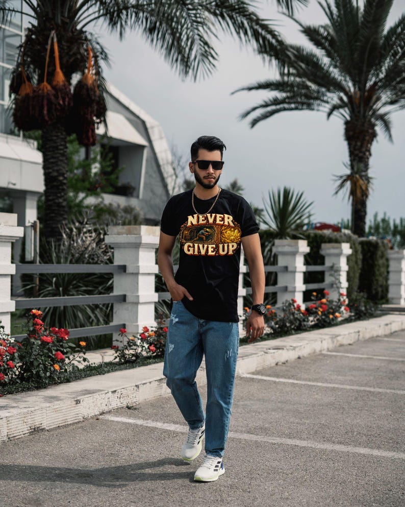 Puede incluir: Una camiseta negra de manga corta con el texto NEVER GIVE UP en letras amarillas sobre una ilustraci&oacute;n gr&aacute;fica, combinada con pantalones vaqueros azules y zapatillas deportivas blancas. La camiseta incluye un dise&ntilde;o detallado en tonos naranja y marr&oacute;n.