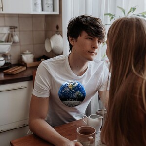 May include: A white short-sleeved t-shirt featuring a circular graphic of the Earth with the text "SAVE ME" printed in white on a dark band across the bottom of the globe. The design is centered on the chest of the garment.
