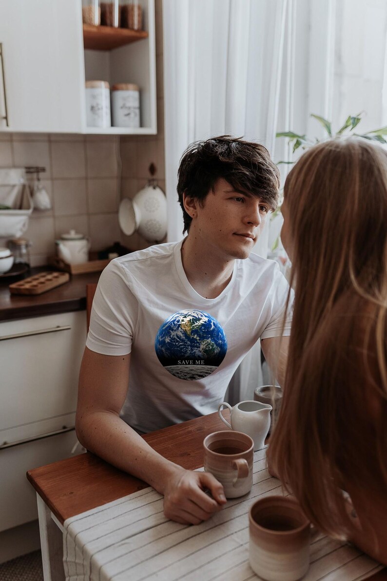 May include: A white short-sleeved t-shirt featuring a circular graphic of the Earth with the text "SAVE ME" printed in white on a dark band across the bottom of the globe. The design is centered on the chest of the garment.