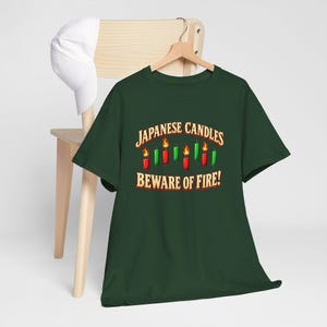May include: A dark green short-sleeve t-shirt featuring a graphic of red and green Japanese candlestick chart bars with flames on top. The text reads "JAPANESE CANDLES BEWARE OF FIRE!" in a bold, light-coloured font centred on the chest.