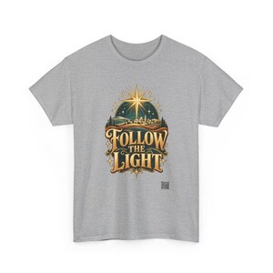 May include: A heather gray short-sleeve t-shirt featuring a central graphic of a glowing gold star above a landscape with the text FOLLOW THE LIGHT in bold gold lettering. The design includes a dark teal sky and rolling hills.