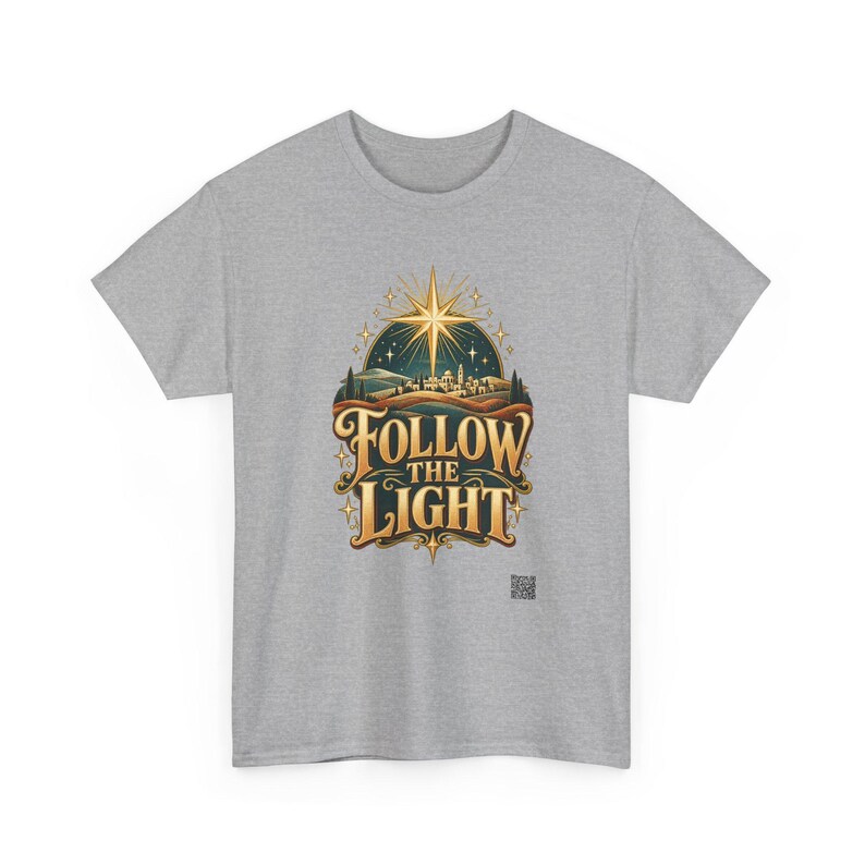 May include: A heather gray short-sleeve t-shirt featuring a central graphic of a glowing gold star above a landscape with the text FOLLOW THE LIGHT in bold gold lettering. The design includes a dark teal sky and rolling hills.