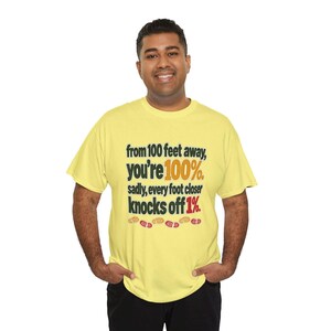 May include: A light yellow short-sleeved t-shirt featuring the black text: from 100 feet away, you're 100%. sadly, every foot closer knocks off 1%. The text includes orange and red accents, with small red and orange footprint icons below.