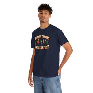 May include: A navy blue t-shirt featuring a graphic of red and green Japanese candlestick chart icons with flames on top. The text reads "JAPANESE CANDLES" above the icons and "BEWARE OF FIRE!" below in a bold, light-coloured font.