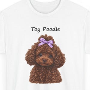 May include: A white t-shirt featuring a graphic illustration of a brown curly-haired toy poodle with a purple bow on its head. The text "Toy Poodle" is printed in black above the dog illustration, which shows the head and chest of the animal.