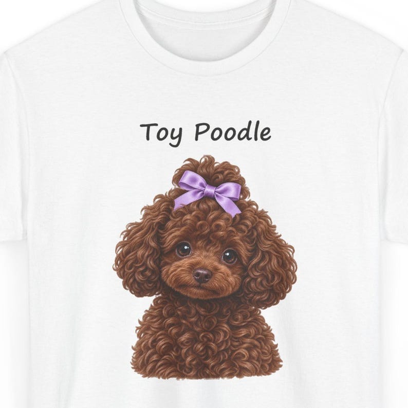 May include: A white t-shirt featuring a graphic illustration of a brown curly-haired toy poodle with a purple bow on its head. The text "Toy Poodle" is printed in black above the dog illustration, which shows the head and chest of the animal.