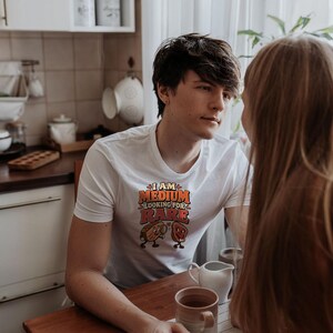 May include: A white short-sleeved t-shirt featuring a graphic print with the text "I AM MEDIUM LOOKING FOR RARE" in orange and red letters, accompanied by illustrations of a medium-cooked steak and a rare-cooked steak with faces.