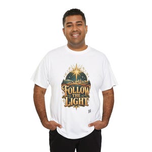 May include: A white short-sleeved t-shirt featuring a graphic print of a glowing star above a landscape with the text FOLLOW THE LIGHT in gold and dark brown lettering. The design includes stylized hills, trees, and small stars.