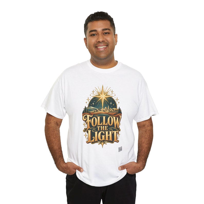 May include: A white short-sleeved t-shirt featuring a graphic print of a glowing star above a landscape with the text FOLLOW THE LIGHT in gold and dark brown lettering. The design includes stylized hills, trees, and small stars.