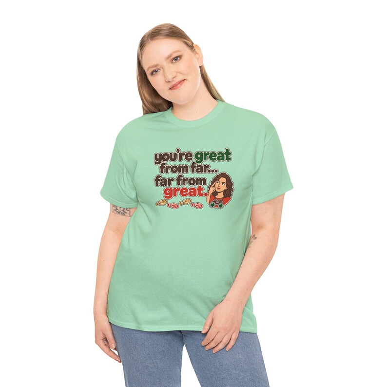 May include: A light green short-sleeve t-shirt featuring the text "you're great from far... far from great." in brown and red lettering, accompanied by a cartoon illustration of a person holding a game controller.