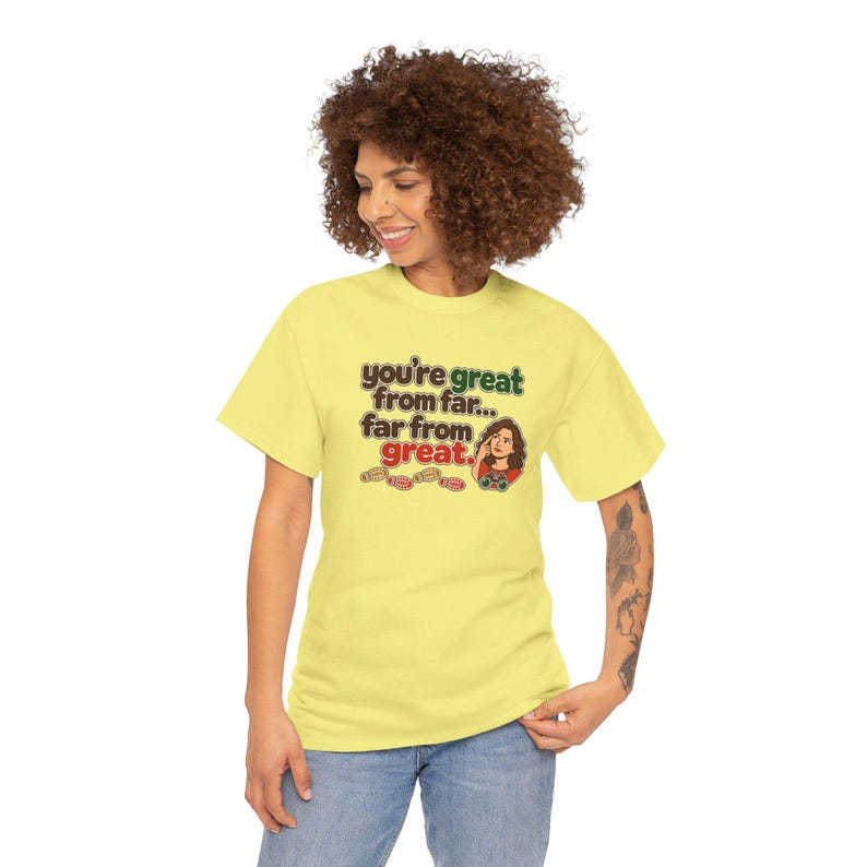 May include: A light yellow short-sleeve t-shirt featuring the brown and red text "you're great from far... far from great." The design includes a small illustration of a person looking through binoculars and several small brown footprint icons.