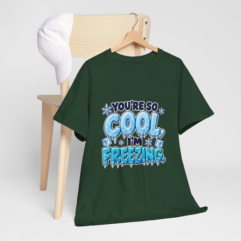 May include: A dark green short-sleeve t-shirt featuring a graphic with the text "YOU'RE SO COOL, I'M FREEZING." The letters are designed to look like blue ice with icicles, surrounded by small white snowflakes and ice cubes.