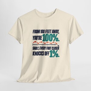 May include: A cream-colored short-sleeve t-shirt featuring dark blue and teal text that reads: FROM 100 FEET AWAY, YOU'RE 100%. SADLY, EVERY FOOT CLOSER KNOCKS OFF 1%. A graphic of small orange footprints follows a horizontal line.