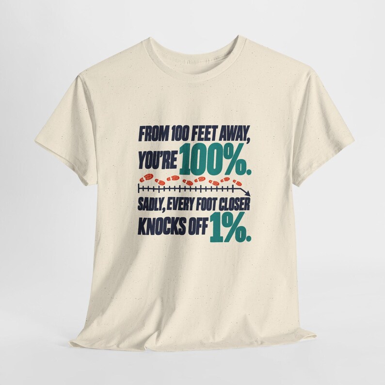 May include: A cream-colored short-sleeve t-shirt featuring dark blue and teal text that reads: FROM 100 FEET AWAY, YOU'RE 100%. SADLY, EVERY FOOT CLOSER KNOCKS OFF 1%. A graphic of small orange footprints follows a horizontal line.