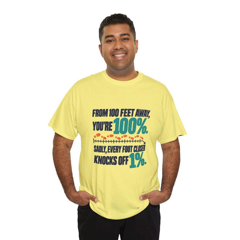 May include: A light yellow short-sleeve t-shirt featuring dark blue text that reads: FROM 100 FEET AWAY, YOU'RE 100%. SADLY, EVERY FOOT CLOSER KNOCKS OFF 1%. The design includes a graphic of orange footprints on a black measurement line.