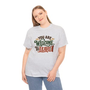 May include: A light gray short-sleeve t-shirt featuring the text "YOU ARE WELCOME... TO LEAVE!" printed on the front. The word "YOU ARE" is in brown, "WELCOME" is in dark green, and "TO LEAVE!" is in red with a small door icon.