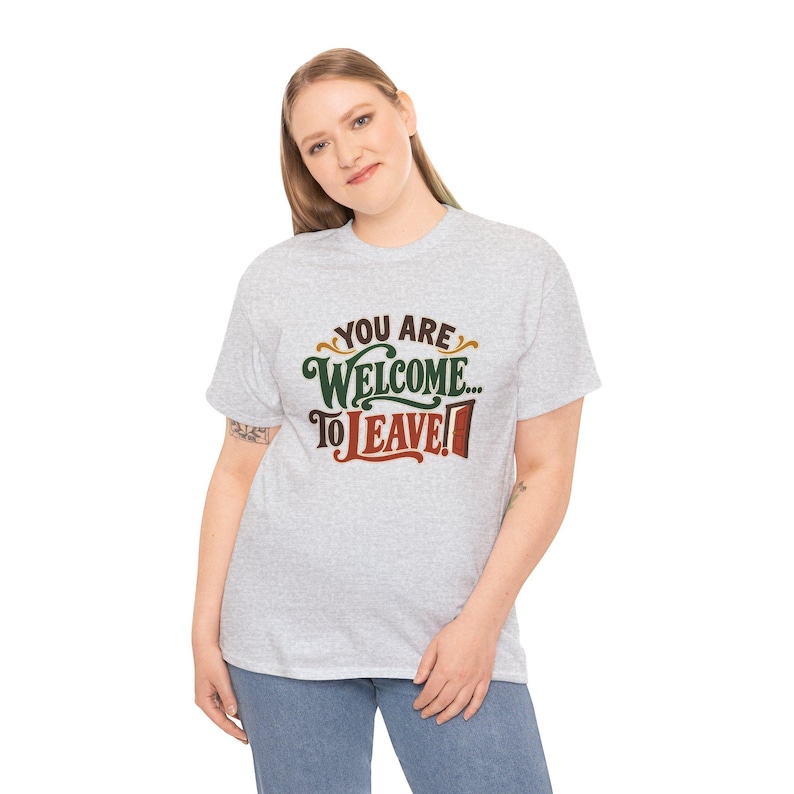 May include: A light gray short-sleeve t-shirt featuring the text "YOU ARE WELCOME... TO LEAVE!" printed on the front. The word "YOU ARE" is in brown, "WELCOME" is in dark green, and "TO LEAVE!" is in red with a small door icon.