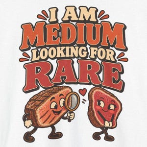 May include: A white t-shirt featuring a graphic print with the text I AM MEDIUM LOOKING FOR RARE in orange and red retro-style lettering. The design includes two cartoon steak characters, one holding a magnifying glass, with a small red heart.