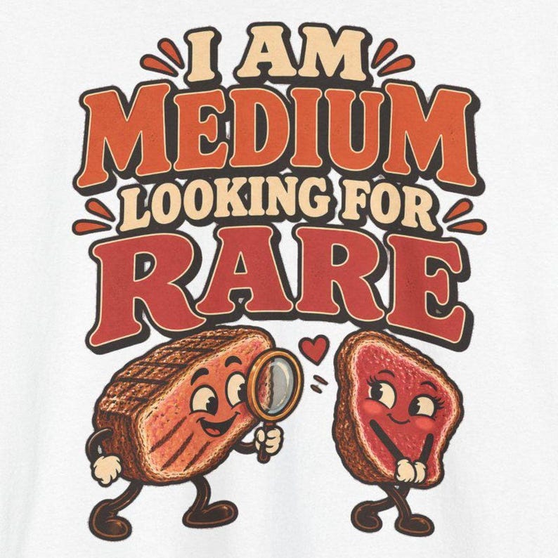 May include: A white t-shirt featuring a graphic print with the text I AM MEDIUM LOOKING FOR RARE in orange and red retro-style lettering. The design includes two cartoon steak characters, one holding a magnifying glass, with a small red heart.