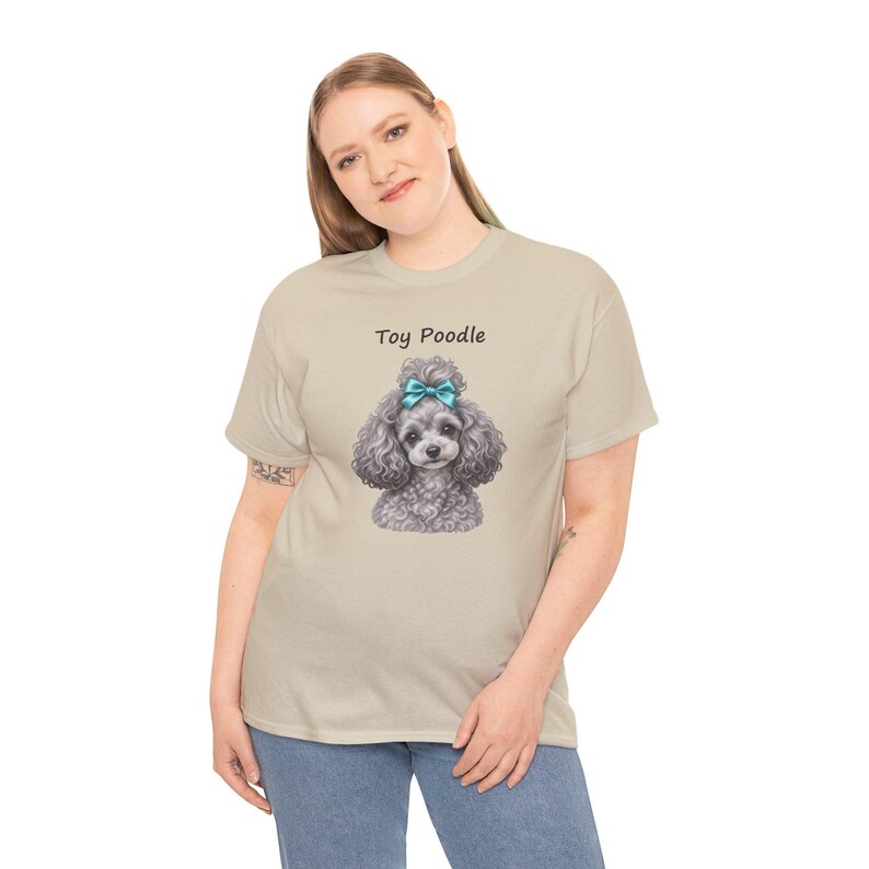 K&ouml;nnte beinhalten: Ein beiges Kurzarm-T-Shirt mit einer Grafik eines grauen Toypudels, der eine leuchtend blaue Schleife auf dem Kopf tr&auml;gt. Der Text Toy Poodle ist in Schwarz &uuml;ber der Hundeillustration gedruckt. Das Shirt hat einen Rundhalsausschnitt.