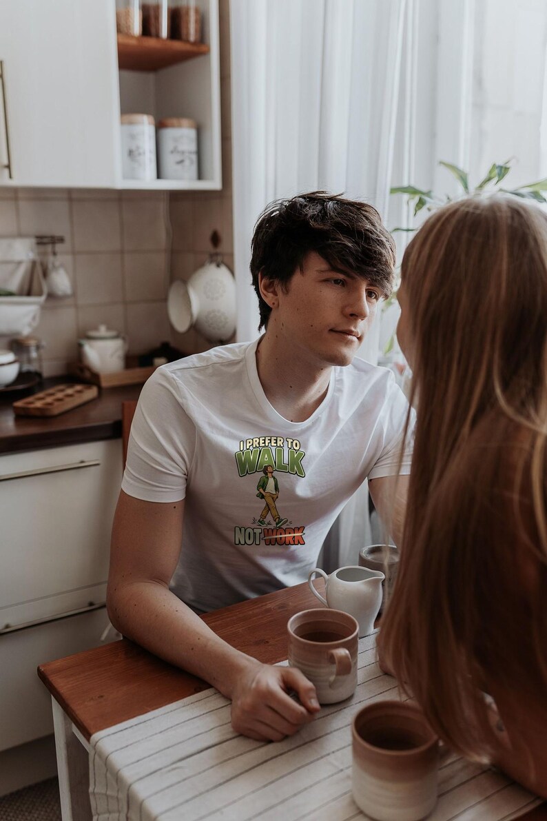 Op de afbeelding: Een wit T-shirt met korte mouwen en een grafische print met de tekst I PREFER TO WALK NOT WORK in groene en rode letters, vergezeld van een illustratie van een persoon die loopt in een groen shirt en een beige broek.