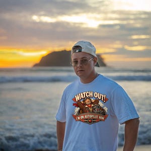 May include: A white short-sleeved t-shirt featuring a graphic of a pilot in a cockpit with a robot. The text reads "WATCH OUT!" at the top and "I'M ON AUTOPILOT NOW." at the bottom, accented with red wings and a propeller icon.