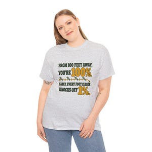 May include: A light gray short-sleeve t-shirt featuring black and gold text that reads: FROM 100 FEET AWAY, YOU'RE 100%. SADLY, EVERY FOOT CLOSER KNOCKS OFF 1%. The design includes a graphic of a measuring line with small foot icons.