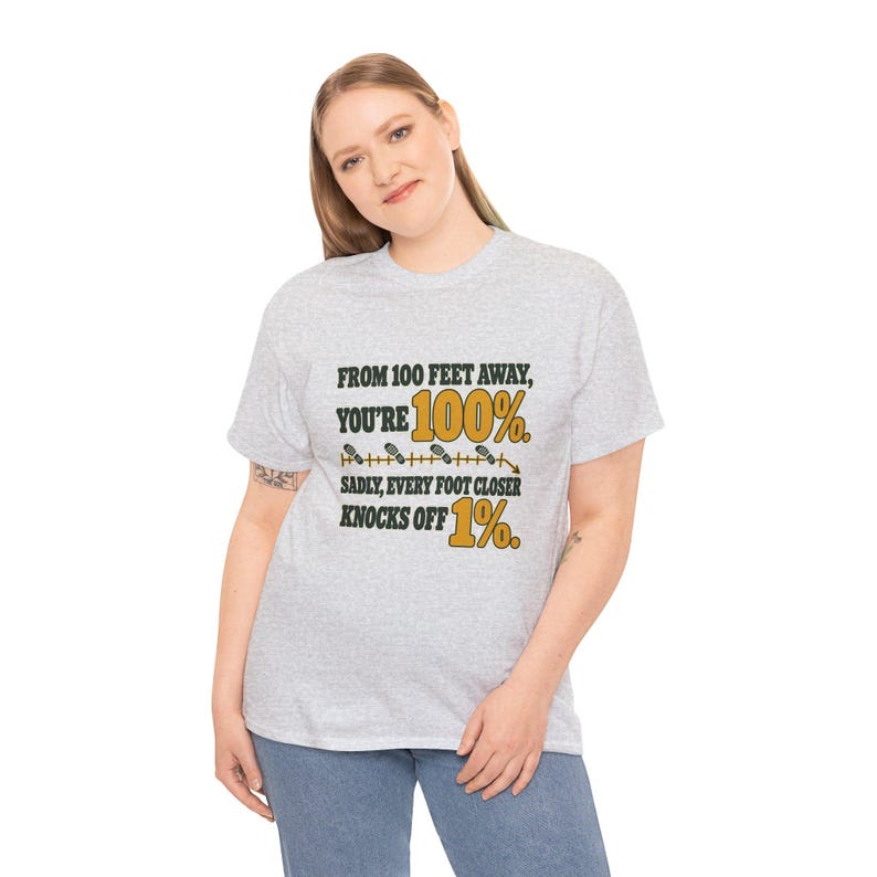 May include: A light gray short-sleeve t-shirt featuring black and gold text that reads: FROM 100 FEET AWAY, YOU'RE 100%. SADLY, EVERY FOOT CLOSER KNOCKS OFF 1%. The design includes a graphic of a measuring line with small foot icons.