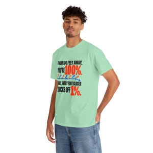 May include: A light green short-sleeve t-shirt featuring black text that reads: FROM 100 FEET AWAY, YOU'RE 100%. AND, EVERY FOOT CLOSER KNOCKS OFF 1%. The design includes a blue arrow graphic with small red and blue footprint icons.