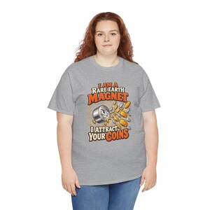 May include: A heather gray short-sleeve t-shirt featuring a graphic of a cartoon magnet with a face, surrounded by gold and silver coins. The text reads, I AM A RARE EARTH MAGNET I ATTRACT YOUR COINS, in bold orange and white lettering.