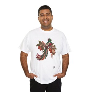 May include: A white short-sleeve t-shirt featuring a central graphic of a green and red quetzal bird perched on a branch with cocoa pods. The design includes intricate geometric patterns in red, white, and brown tones.