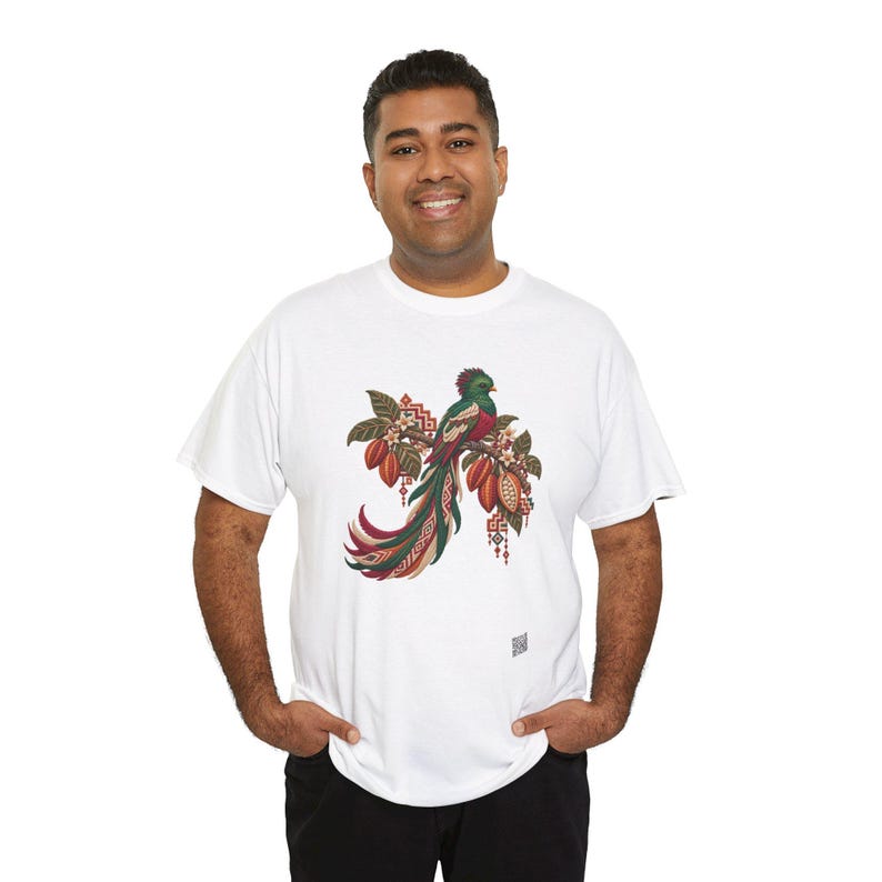 May include: A white short-sleeve t-shirt featuring a central graphic of a green and red quetzal bird perched on a branch with cocoa pods. The design includes intricate geometric patterns in red, white, and brown tones.