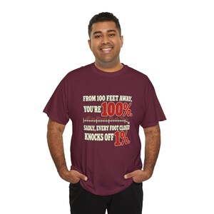May include: A maroon short-sleeve t-shirt featuring the text: FROM 100 FEET AWAY, YOU'RE 100%. SADLY, EVERY FOOT CLOSER KNOCKS OFF 1%. The design includes a horizontal line graphic with small brown shapes and red percentage numbers.