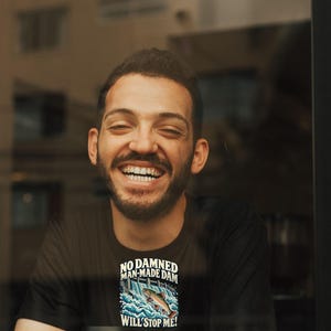Puede incluir: Una camiseta negra con un gr&aacute;fico de un pez saltando sobre una presa y el texto "NO DAMNED MAN-MADE DAM WILL STOP ME!" impreso en blanco y azul, junto a un vaso de caf&eacute; desechable blanco con tapa negra y un logotipo negro.