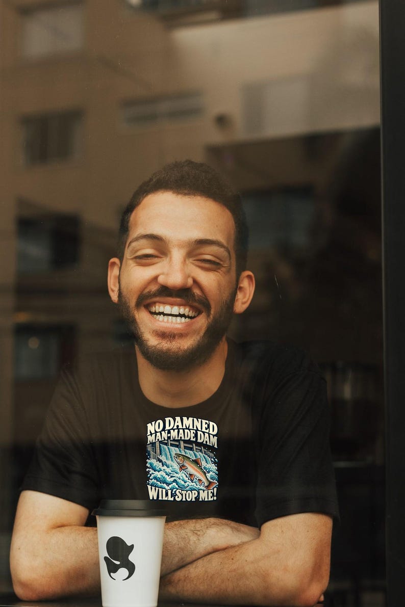 Puede incluir: Una camiseta negra con un gr&aacute;fico de un pez saltando sobre una presa y el texto "NO DAMNED MAN-MADE DAM WILL STOP ME!" impreso en blanco y azul, junto a un vaso de caf&eacute; desechable blanco con tapa negra y un logotipo negro.