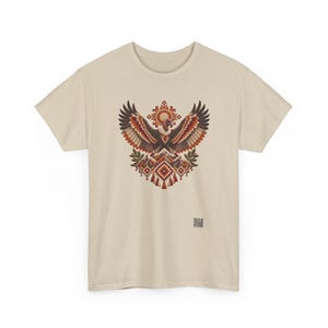 May include: A beige short-sleeve t-shirt featuring a central graphic of a vulture with outstretched wings above a mountain range, accented by geometric patterns in shades of red, brown, and cream, with a small QR code printed near the bottom right.
