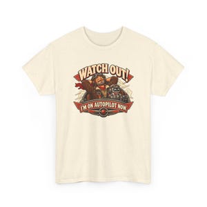 May include: A cream-colored t-shirt featuring a graphic of a pilot and a robot in a cockpit. The text reads WATCH OUT! I'M ON AUTOPILOT NOW. The design includes red wings, a propeller, and clouds in a brown, red, and white color palette.