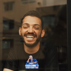May include: A black t-shirt featuring a graphic of a man with grey hair resting his head on his hand above a chat interface that reads HI. Ask me anything, with icons for new chat, voice, and image, positioned above a white paper cup.