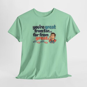 May include: A light green t-shirt featuring a graphic with the text "you're great from far... far from great." The design includes a cartoon character wearing binoculars and pointing to their head, accompanied by small red and blue footprint icons.