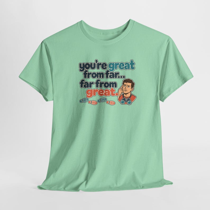 May include: A light green t-shirt featuring a graphic with the text "you're great from far... far from great." The design includes a cartoon character wearing binoculars and pointing to their head, accompanied by small red and blue footprint icons.