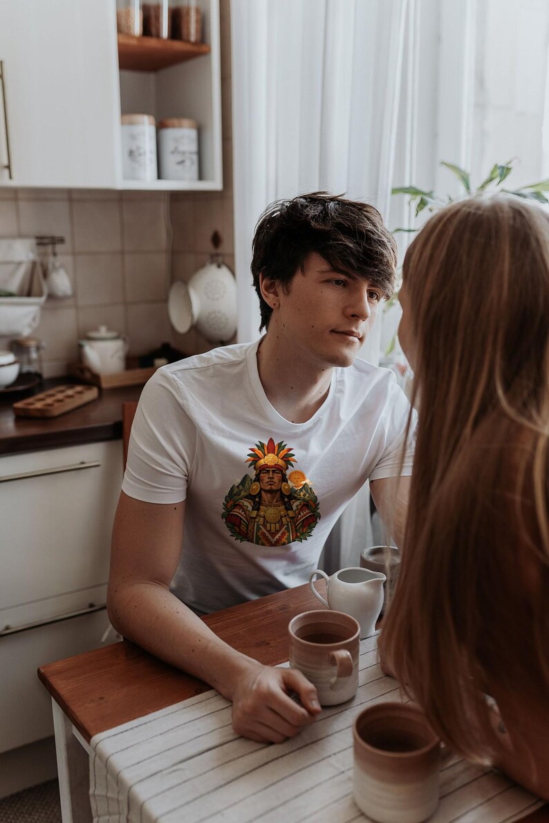 May include: A white short-sleeved crew neck t-shirt featuring a colorful graphic print on the chest. The design depicts a figure wearing an elaborate feathered headdress and traditional attire in shades of red, orange, yellow, and green.