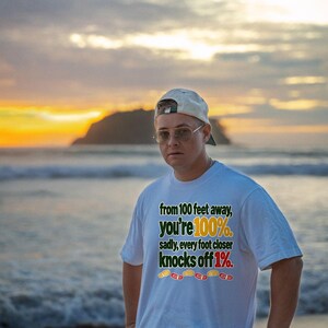 May include: A white short-sleeved t-shirt featuring the text: from 100 feet away, you're 100%. sadly, every foot closer knocks off 1%. The text is accompanied by a row of five small, colorful shoe print icons in yellow, red, and orange.